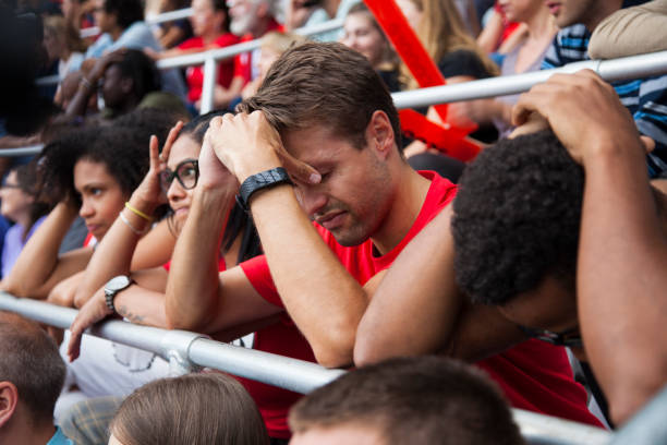 A man in a red shirt with his head in his hands disappointed surrounded by other people sitting disappointed