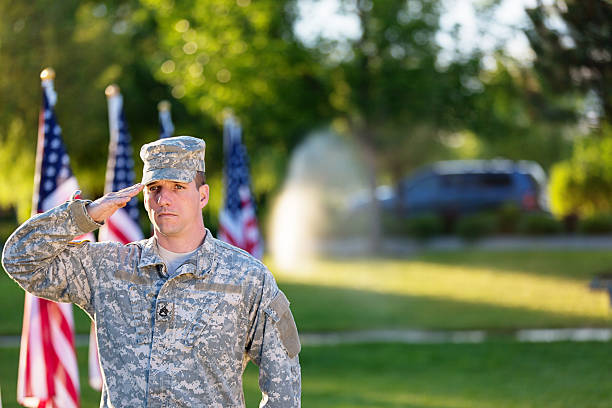 A man in an army unfiorm standing at attention outside with a flag in the background
