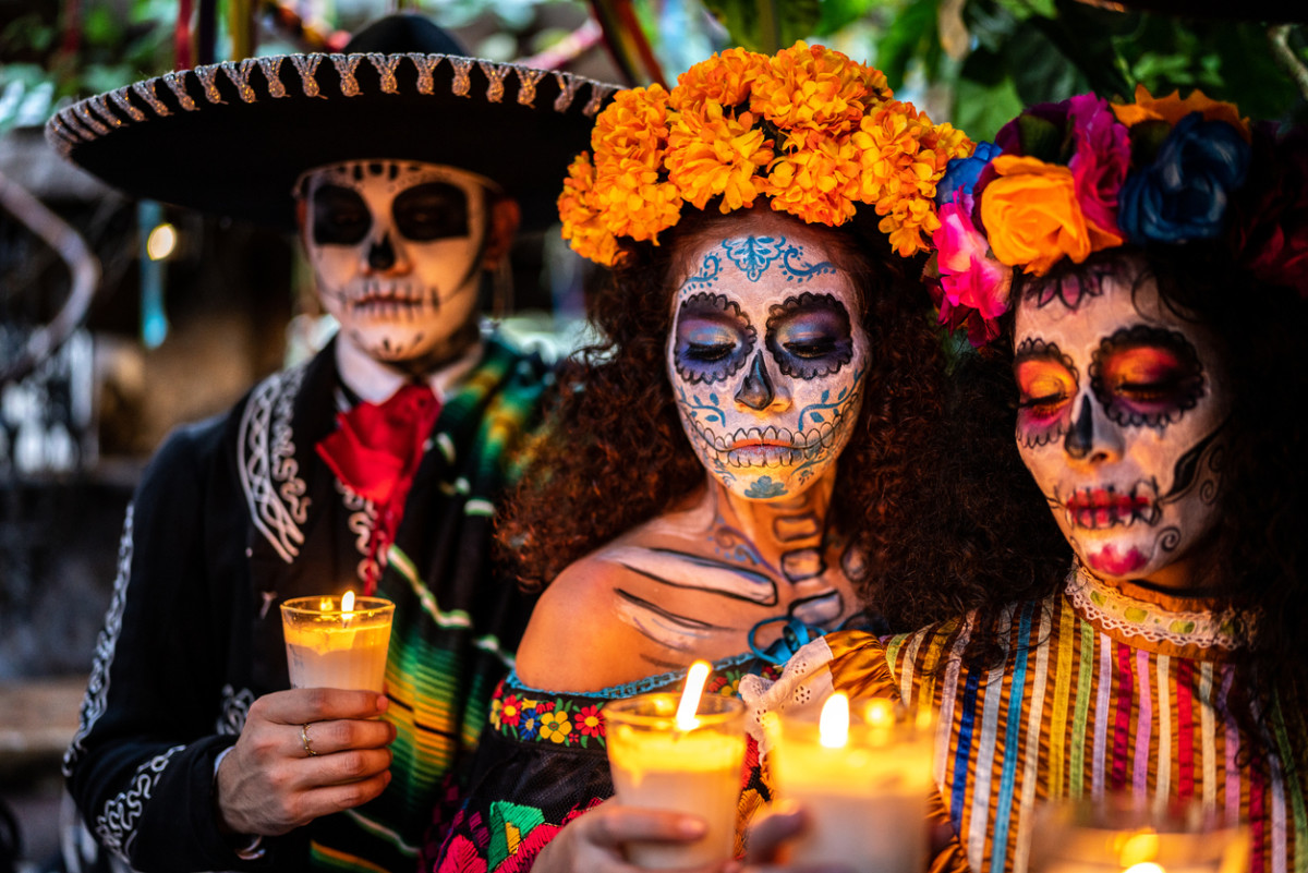 Three people with painted faces in traditional clothing holding candles for Dia de los Muertos