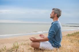 A man meditating by the water in a blue shirt and white shorts