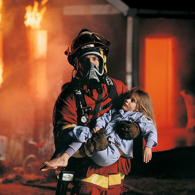 A firefighter in gear holding a child walking away from a burning building