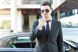 A man in a suit and sunglasses talking into a walkie-talkie ouside a building in front of a car